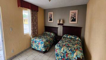 A room with two beds, a window with curtains, a wall-mounted lamp, and framed pictures.