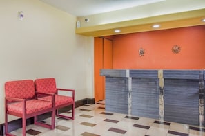 Un área de recepción con mostrador de registro, dos sillas rojas y suelo de baldosas.