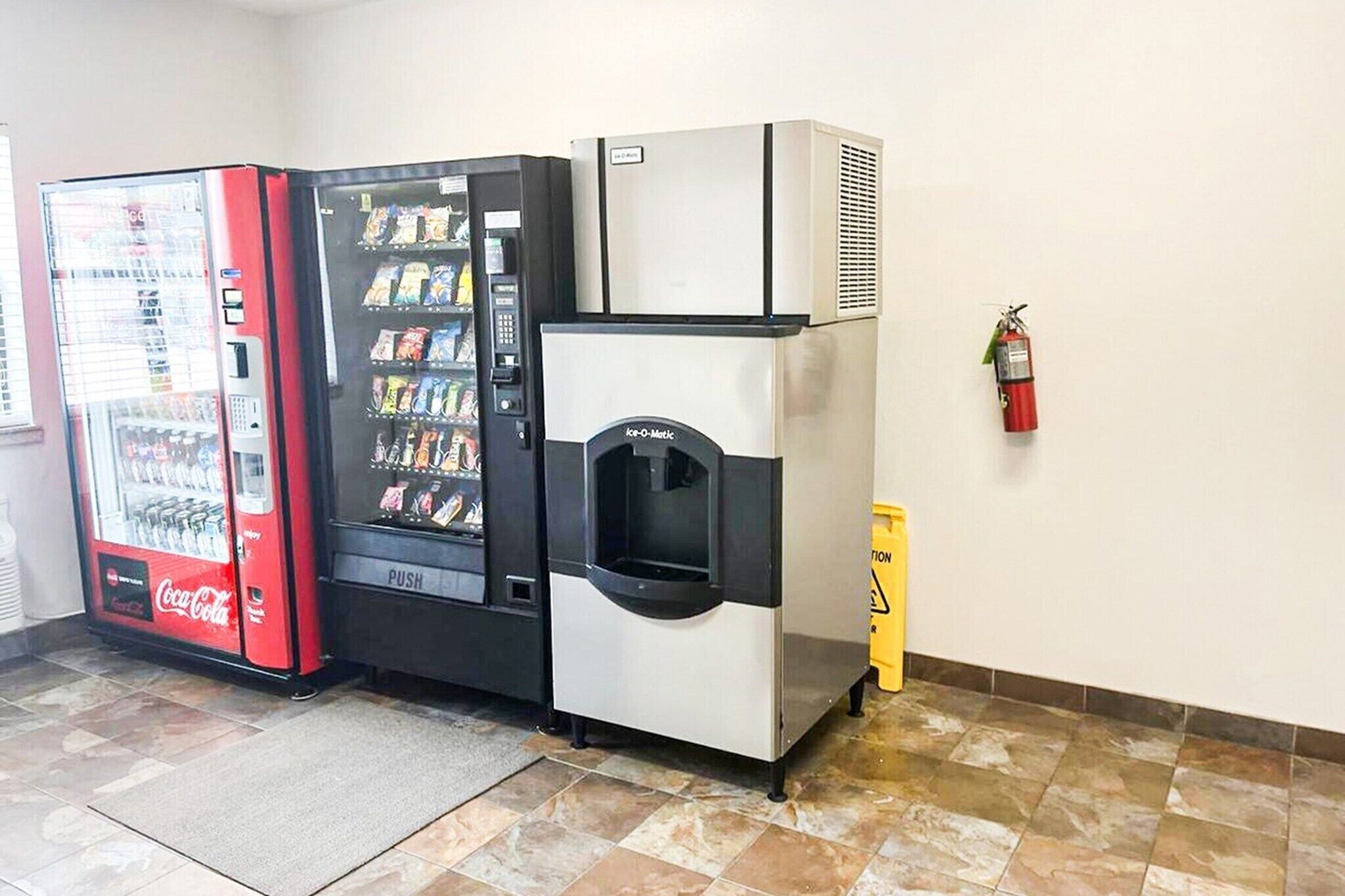 A vending machine, a push-button drink dispenser, and a fire extinguisher in a room with tiled flooring.