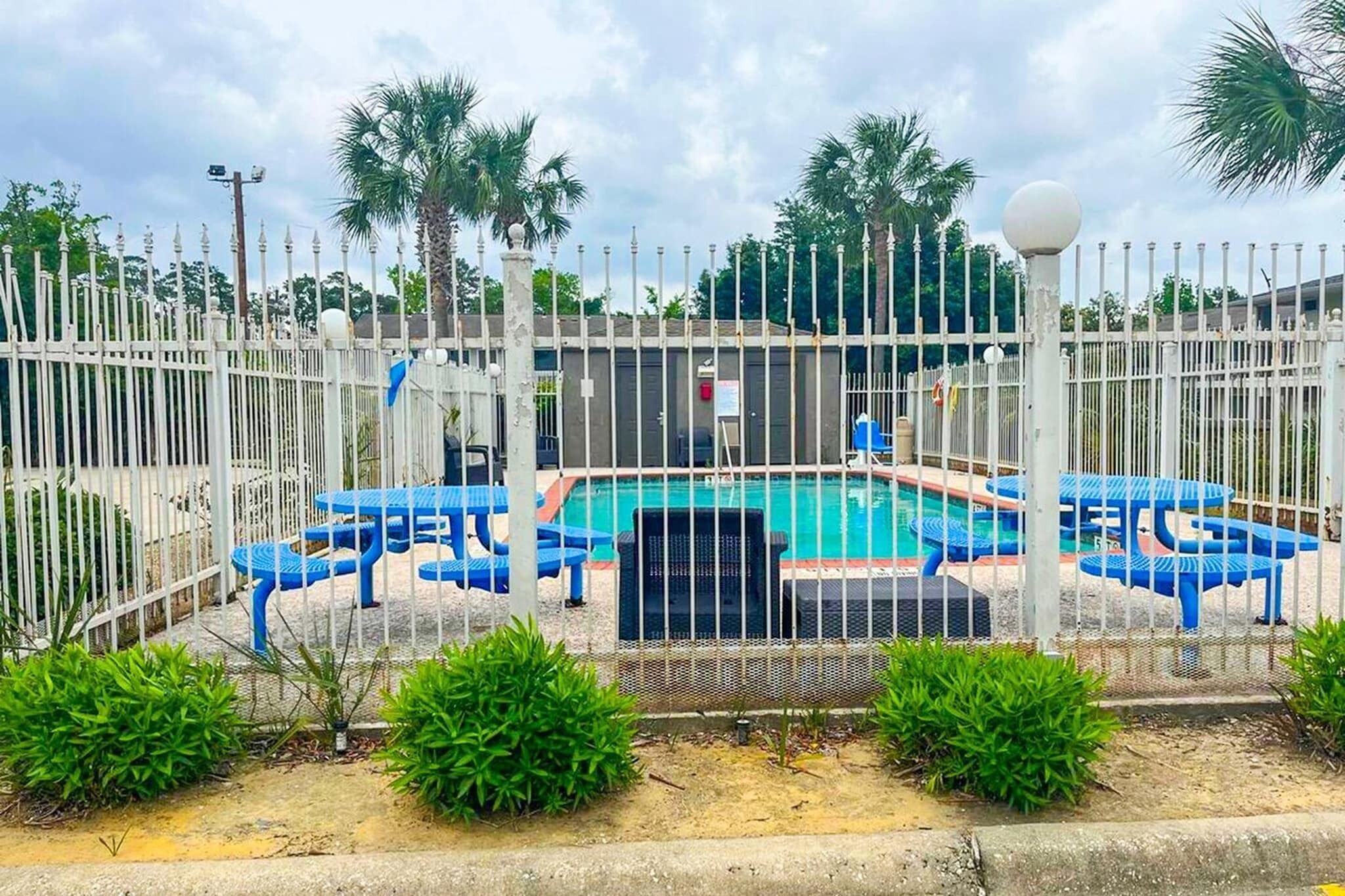 Une piscine entourée d’une clôture, avec des chaises bleues et une petite table, le tout agrémenté de verdure et de palmiers.