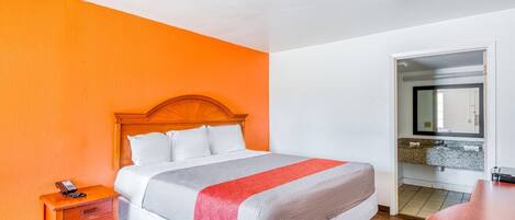 A hotel room with a bed, a nightstand, a desk, a mirror, and a bathroom visible through an open door.