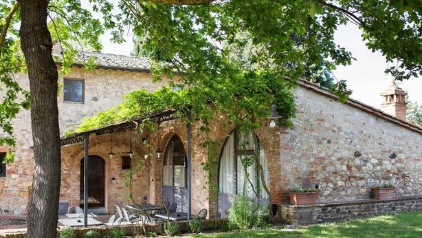 A stone building with a covered patio, outdoor seating, and a tree in the foreground. - Monacianello -Fontebelvedere wine estate (Ponte A Bozzone)