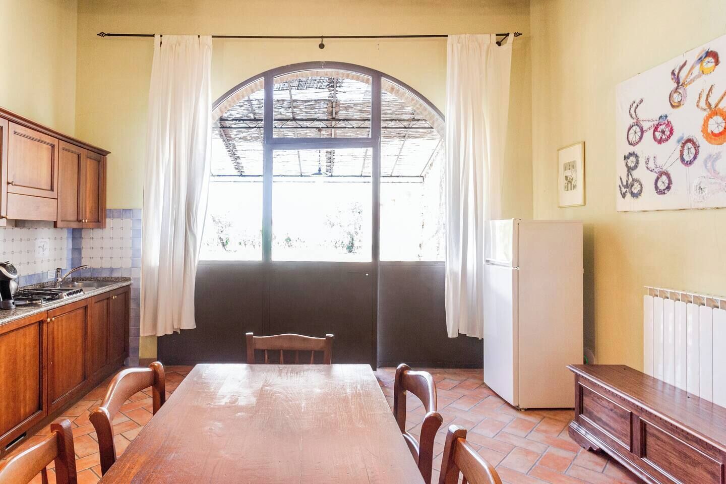 A kitchen with wooden cabinets, a dining table, chairs, a refrigerator, and a large window with curtains.