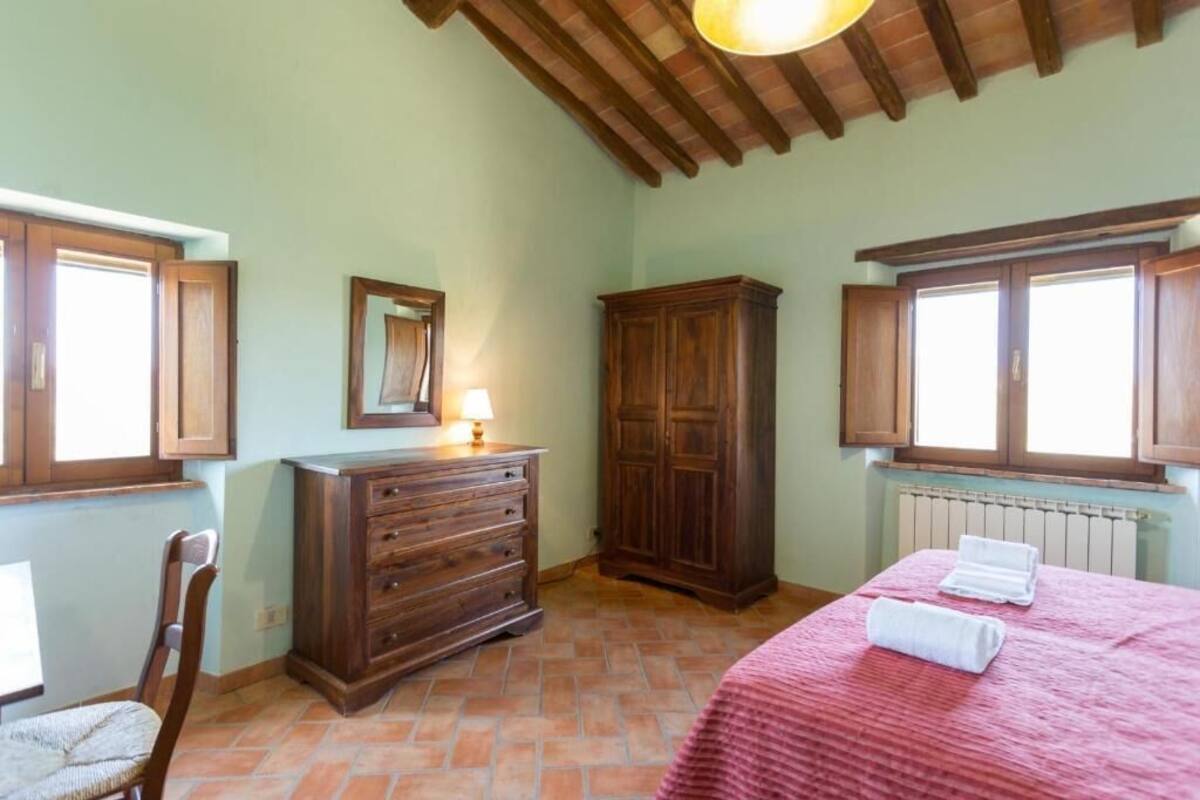 Ein Schlafzimmer mit einem hölzernen Schminktisch, einem Bett mit roter Bettdecke, einem Sessel und einem Fenster mit Holzjalousien.