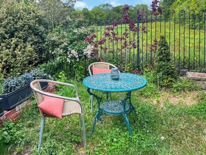 Une terrasse de jardin avec des chaises en osier et une table en métal, entourée d’une végétation luxuriante et d’une clôture en métal.