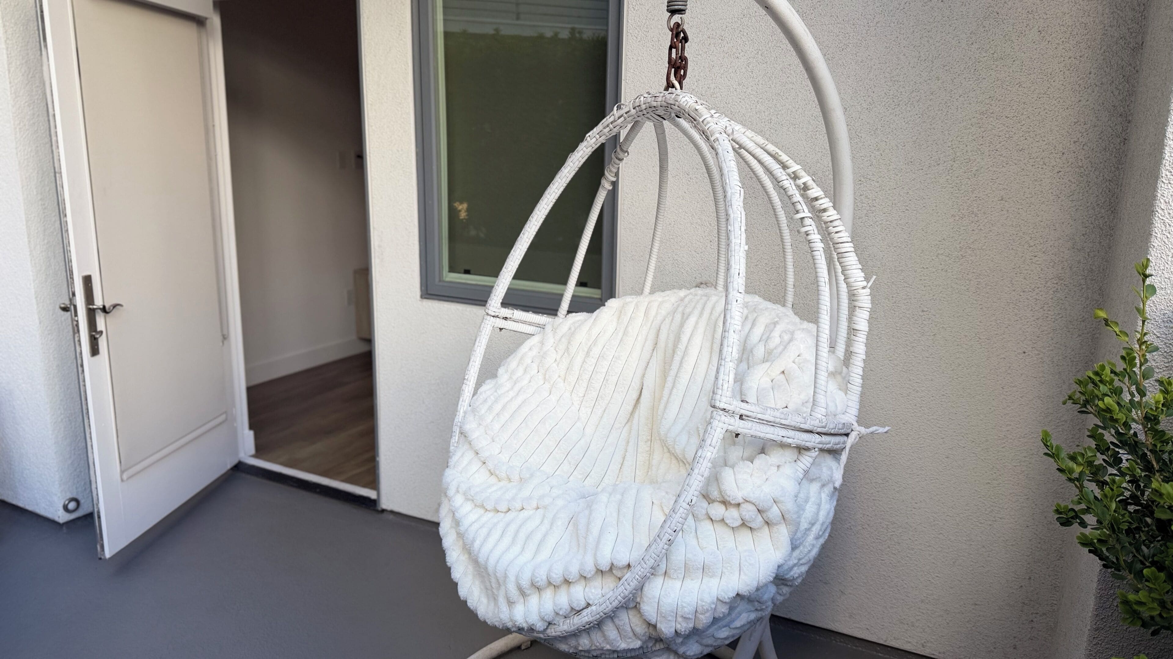 A white wicker hanging chair with a cushion, positioned against a wall near a door and a window.