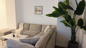 A living room with a beige sectional sofa, a white coffee table, a potted plant, and a framed sign on the wall.