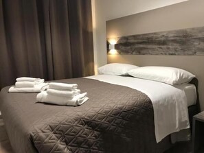Un lit bien fait, recouvert d’une courtepointe matelassée, une lampe fixée au mur et un grand tableau abstrait.