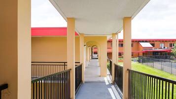 A covered corridor with yellow walls, black railings, and a red-roofed building in the background.
