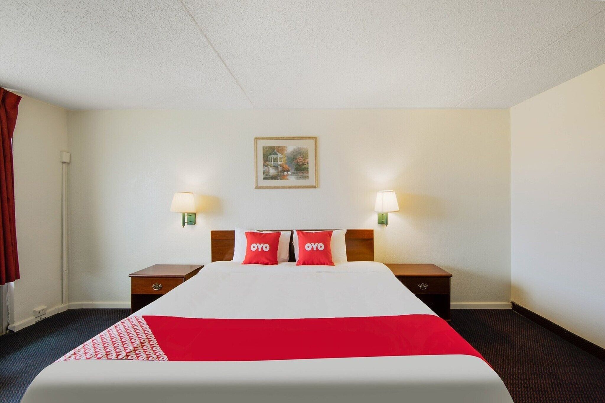 A hotel room with a bed, two bedside tables, two wall-mounted lamps, a framed picture, and a red and white bedspread.