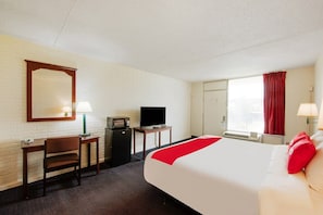 Habitación de hotel con cama, escritorio, silla, televisor, espejo, ventana con cortinas y equipo de aire acondicionado.