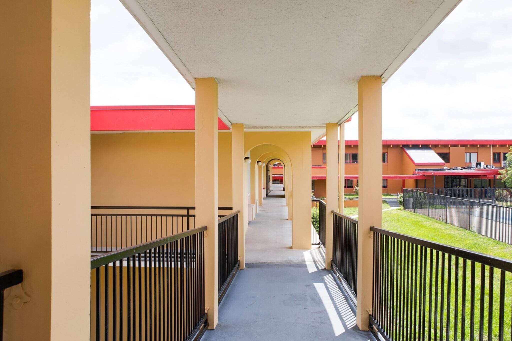 A covered corridor with yellow walls, black railings, and a red-roofed building in the background.