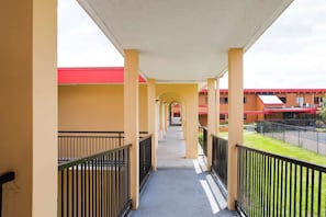 A covered corridor with yellow walls, black railings, and a red-roofed building in the background.