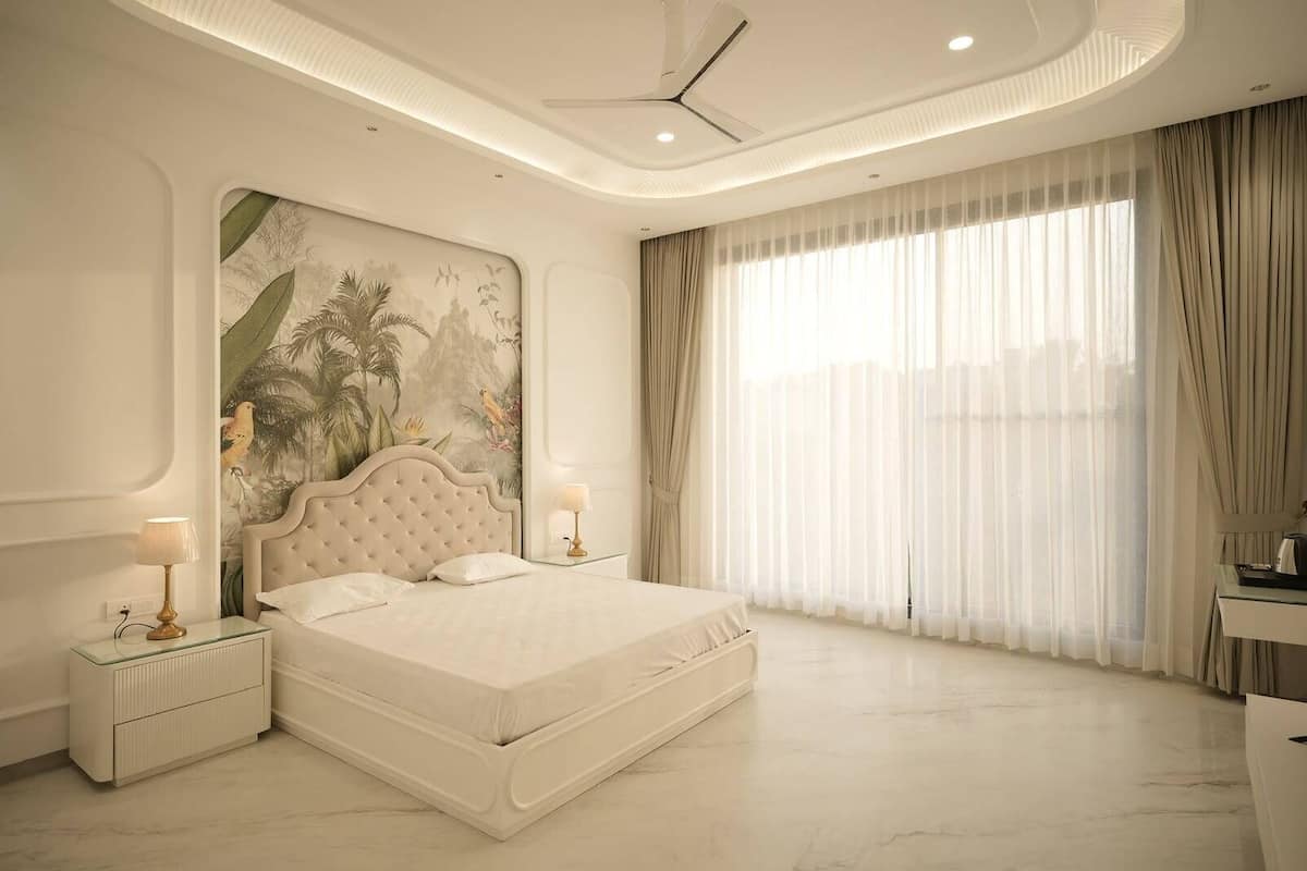 Une chambre moderne avec un grand lit, deux tables de chevet avec des lampes, un ventilateur de plafond et une grande fenĂȘtre avec des rideaux lĂ©gers.