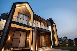 A modern two-story house with a balcony, large windows, and a well-lit entrance.