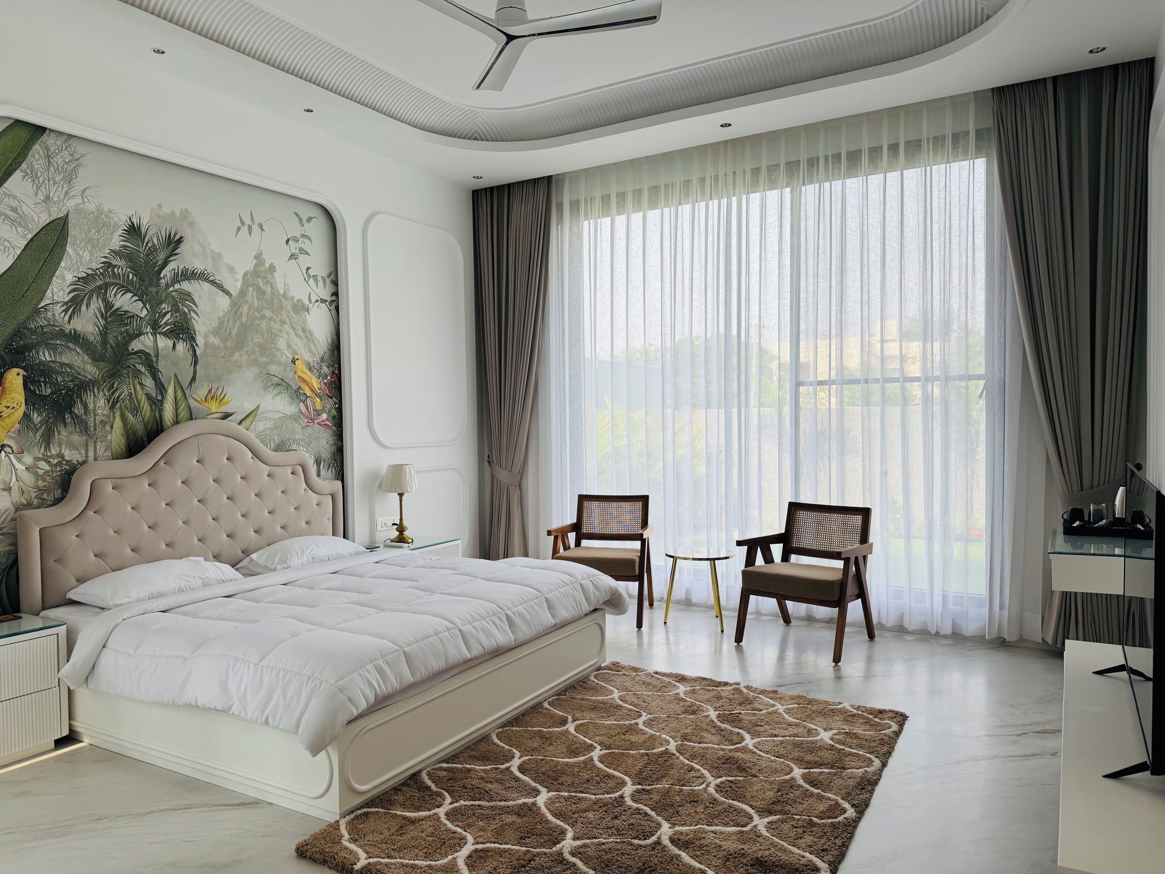 Une chambre à coucher avec un grand lit, deux fauteuils, un bureau et une grande fenêtre avec des rideaux.
