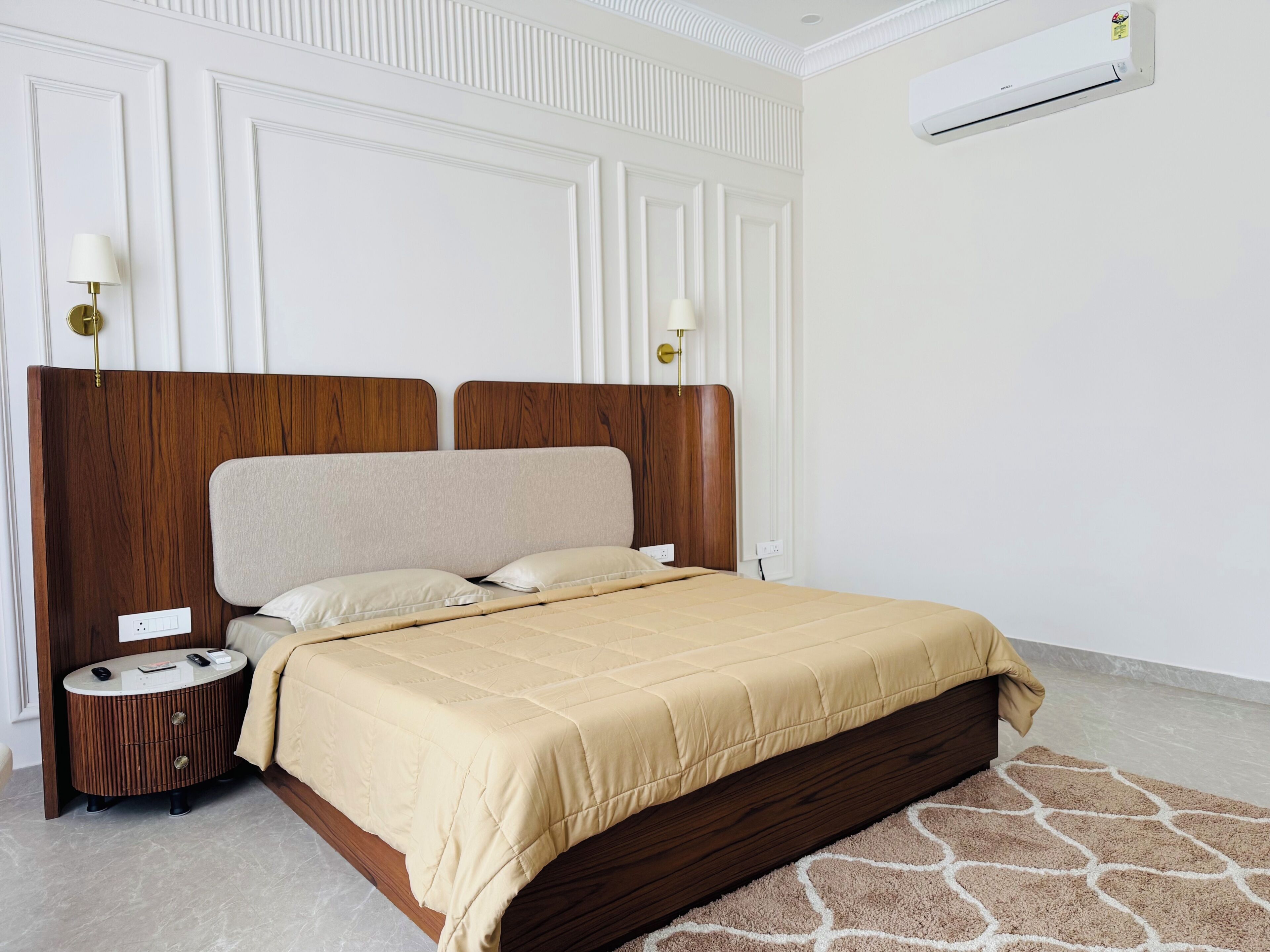 A bedroom with a wooden headboard, a beige bedspread, a round bedside table, and a wall-mounted air conditioner.