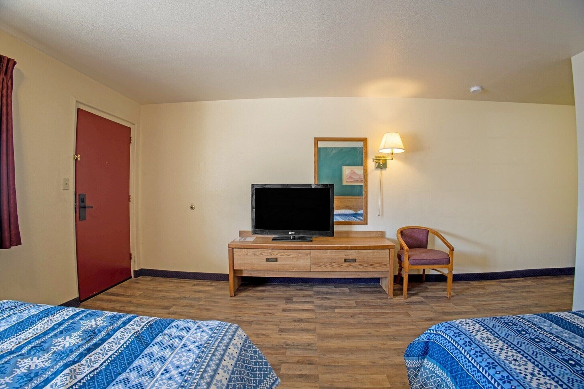 Habitación de hotel con dos camas, un televisor sobre un soporte de madera, una silla y una lámpara fijada a la pared.