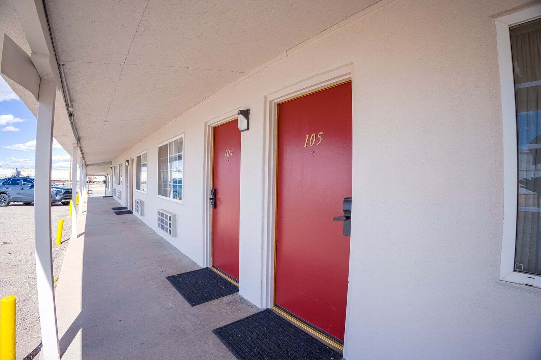 Une rangée de portes rouges, chacune portant un numéro, un passage couvert et un parking.