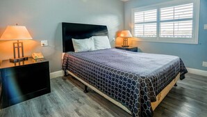 A bedroom with a bed, two bedside tables with lamps, a window with blinds, and a wooden floor.