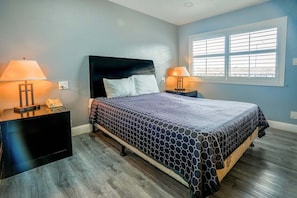 Un dormitorio con una cama, dos mesitas de noche con lámparas, una ventana con persianas y suelo de madera.
