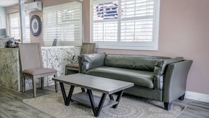 A room with a leather sofa, a wooden coffee table, a patterned rug, a clock on the wall, and a window with blinds. - King Bed Non-Smoking (Buttonwillow, CA)