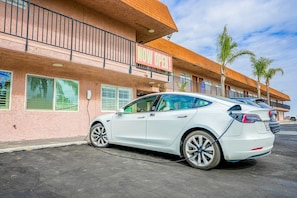 A white car parked in front of a building with a 'NOW OPEN' sign, a balcony, and palm trees.