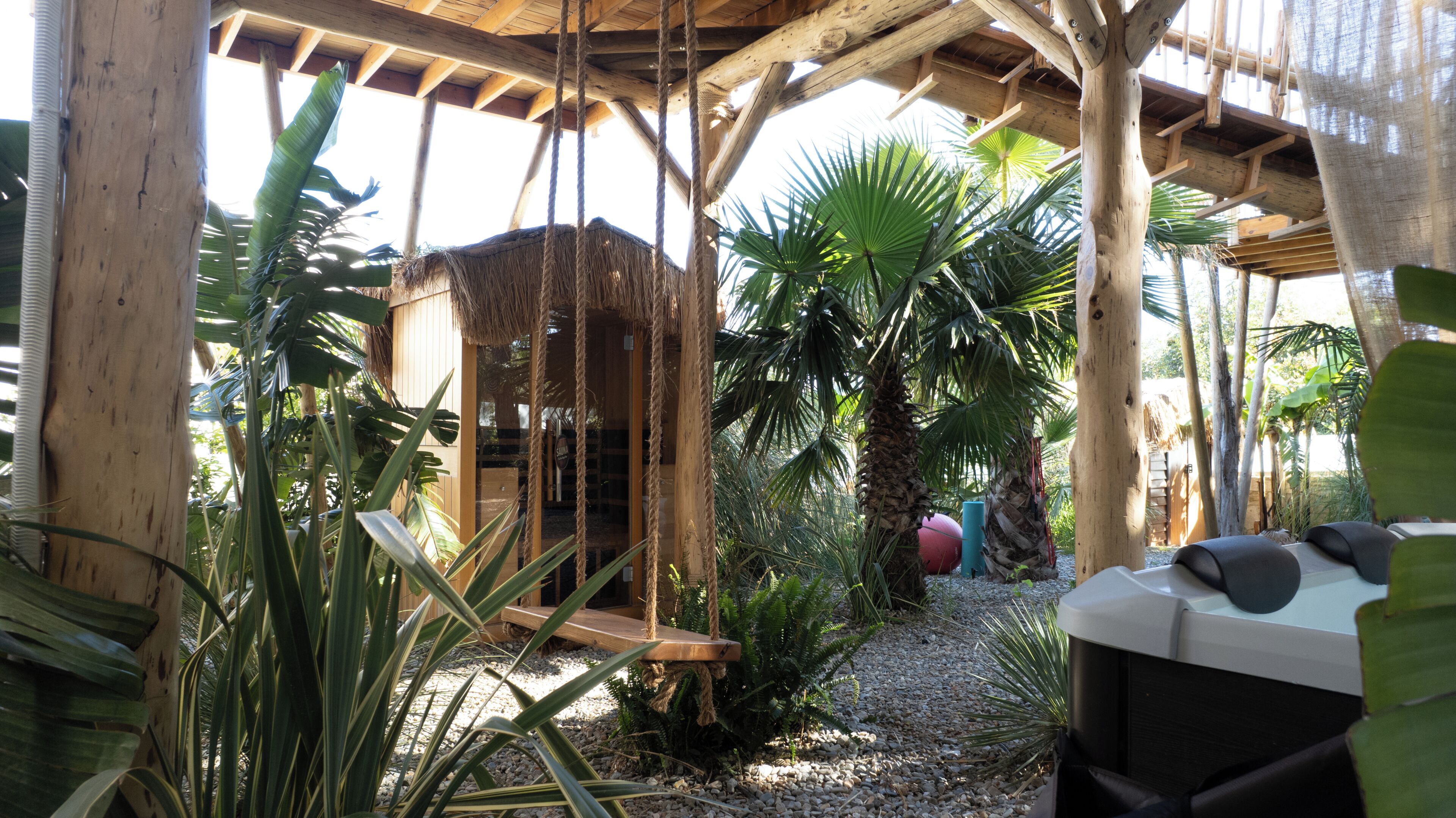 A wooden structure with a thatched roof, surrounded by tropical plants and a gravel path.