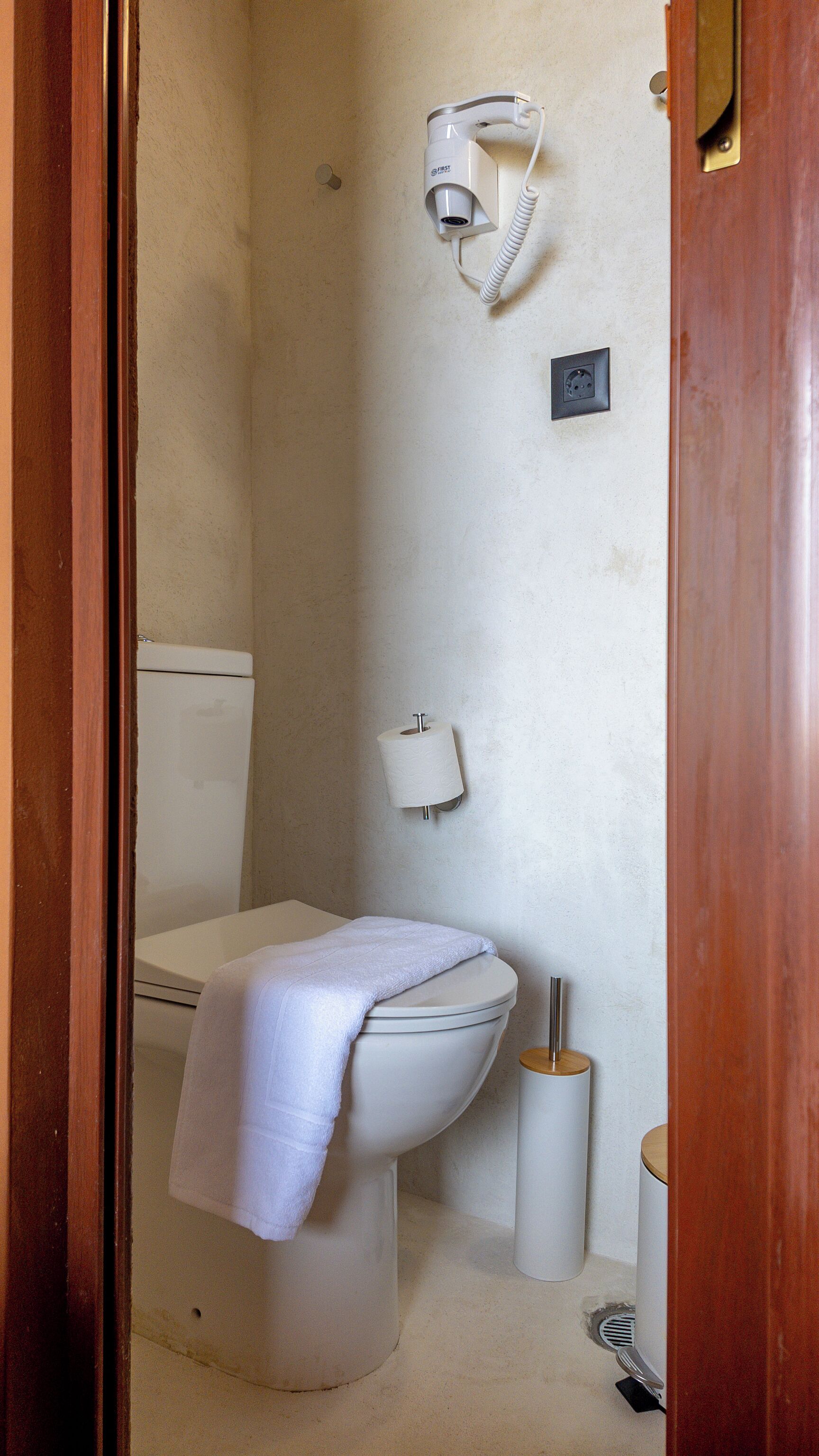 Une salle de bain équipée d’un WC, d’une serviette, d’un porte-rouleau de papier toilette, d’un sèche-cheveux et d’une petite poubelle.