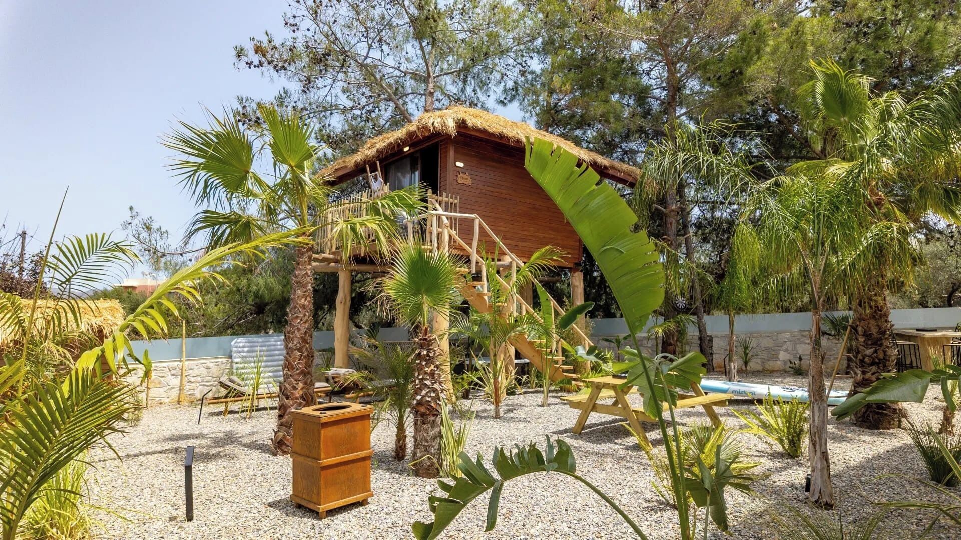 A rustic wooden structure with a thatched roof, surrounded by tropical plants and a gravel path.