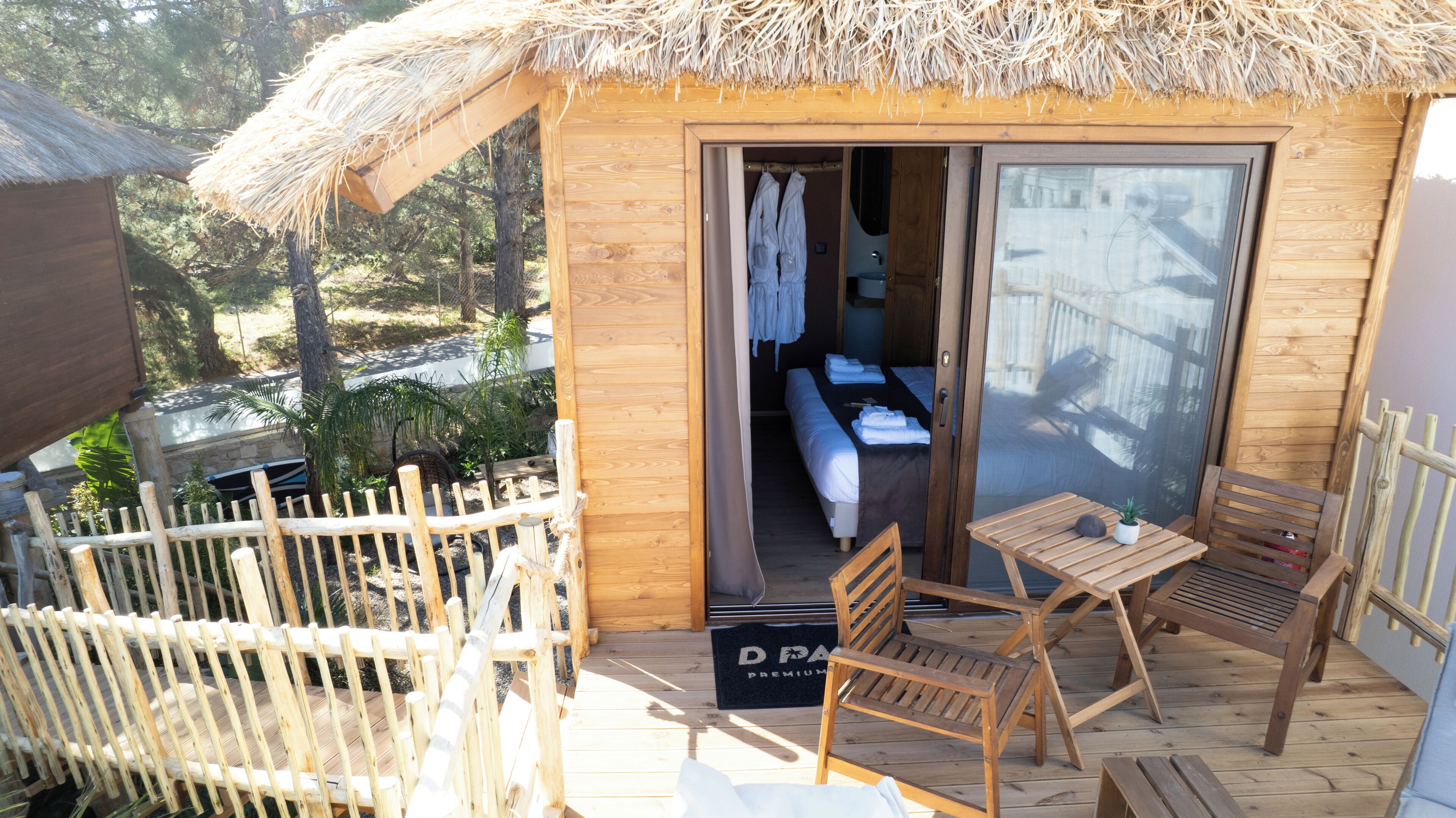 Una cabaña de madera con techo de paja, un balcón con mobiliario de madera y una cama a la vista en el interior.