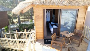 Una cabaña de madera con techo de paja, un balcón con mobiliario de madera y una cama a la vista en el interior.