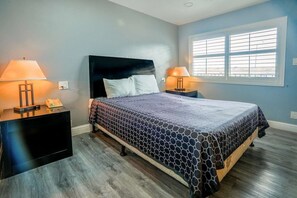 A bedroom with a bed, two bedside tables, a window with blinds, and a wall socket. - Queen Bed Non-Smoking (Buttonwillow, CA)