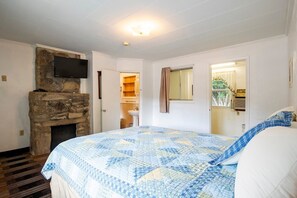 A bedroom with a stone fireplace, a bed with a blue quilt, a television mounted on the wall, and a bathroom visible through an open door. - Queen Room - RiverFront (Bryson city)