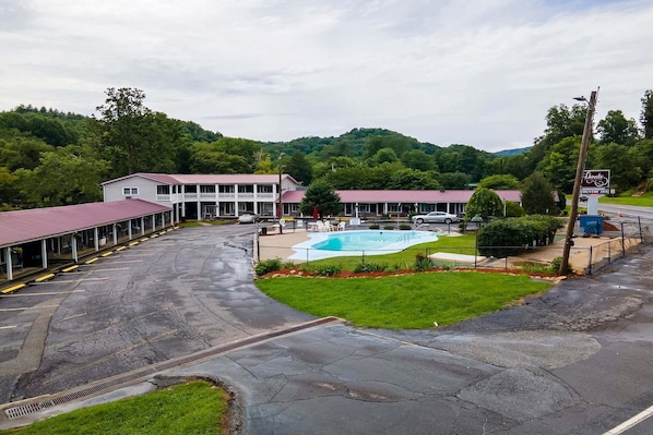 A hotel with a swimming pool, covered parking, and a view of a forested hill.