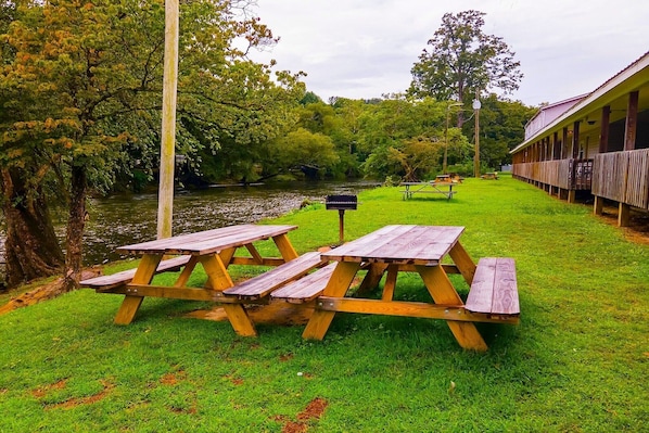 A riverside picnic area with wooden tables and benches, a covered building, and lush greenery.