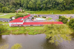 A building with a red roof, adjacent to a body of water, surrounded by greenery and a road.