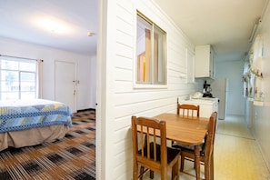 A room with a bed, a wooden dining table with chairs, a kitchen area with cabinets, and a window with curtains.