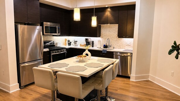 A modern kitchen with dark cabinetry, stainless steel appliances, a dining area with a table and chairs, and a hanging pendant light. - Calm & Stylish Hollywood Gateway (Los Angeles)
