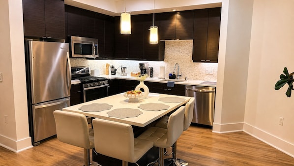 Une cuisine moderne avec des meubles de couleur sombre, des appareils électroménagers en acier inoxydable, un coin repas avec une table et des chaises, et un luminaire suspendu.