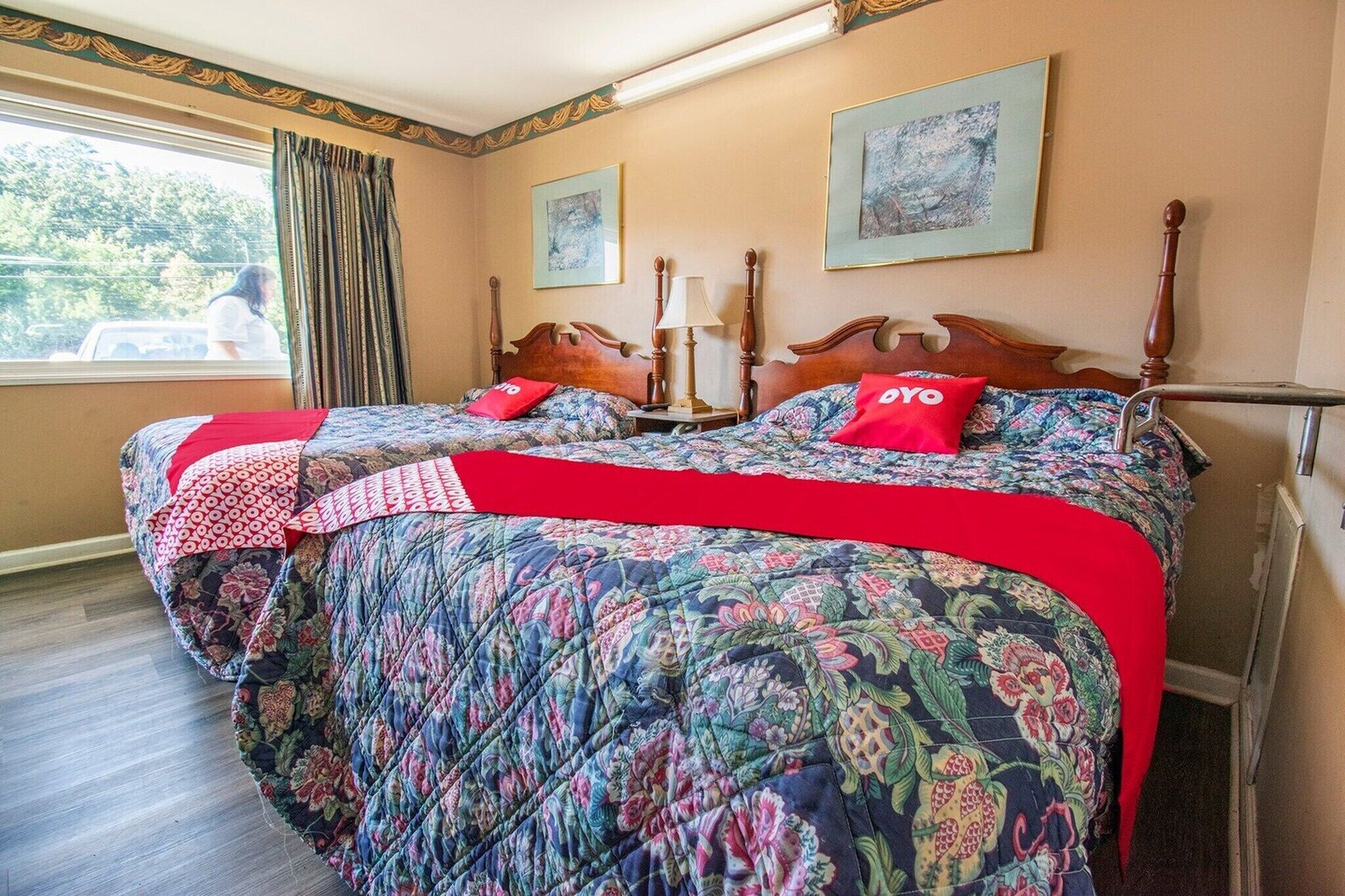 A bedroom with a large bed, two bedside tables, a window with curtains, and two framed artworks on the wall.