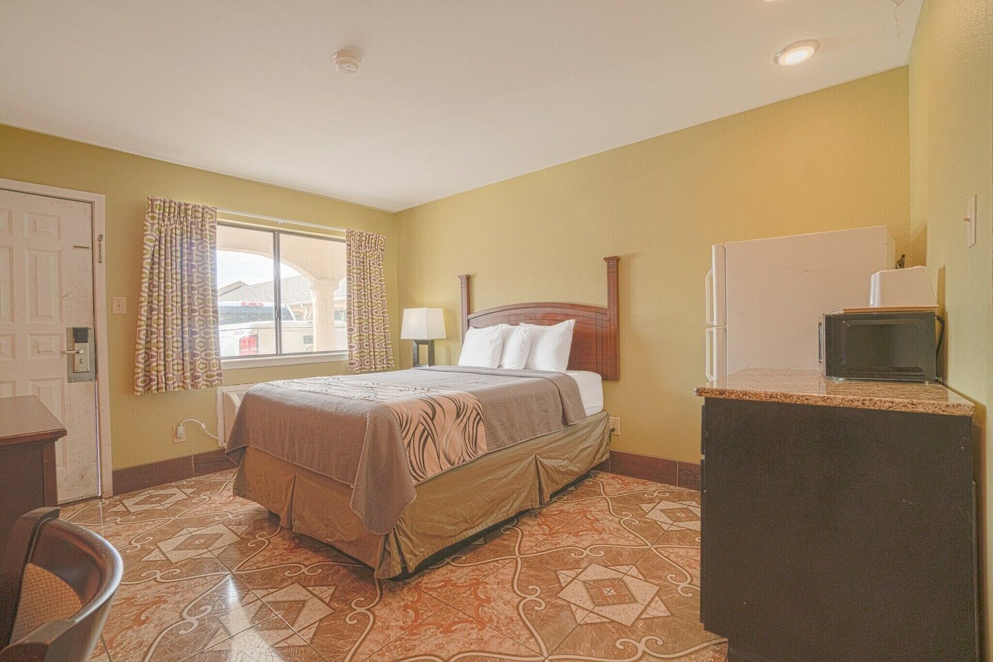 A hotel room with a bed, a desk, a refrigerator, and a window with curtains.
