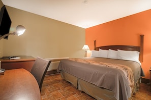 A hotel room with a bed, desk, chair, lamp, and a heater.
