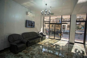A lobby with a chandelier, a sofa, a chair, a framed picture, and a view of the street through large windows. - Hotel Lord (Batumi)