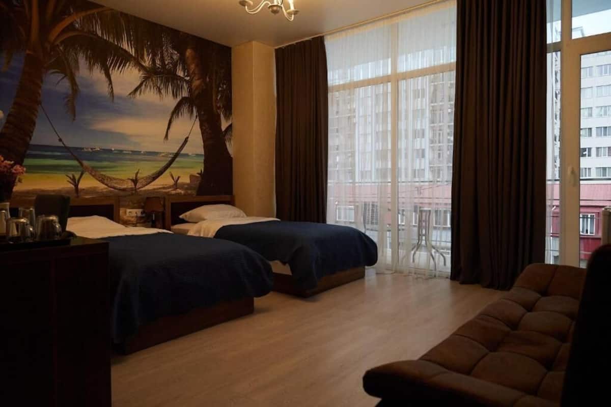 A hotel room with two beds, a sofa, a large window with curtains, and a mural of a beach scene.