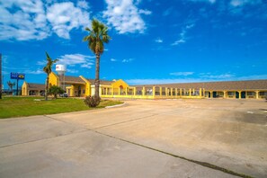 A yellow building with a large parking lot and a palm tree. - Double Queen Room|Corpus Christi (Corpus Christi TX)