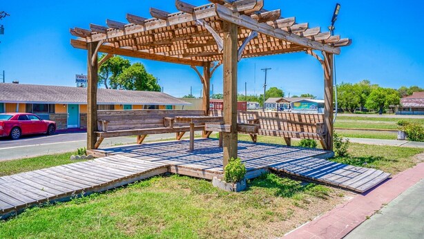 Un kiosque en bois, doté de bancs et d’une petite table, entouré d’une zone pavée et d’un espace herbeux.