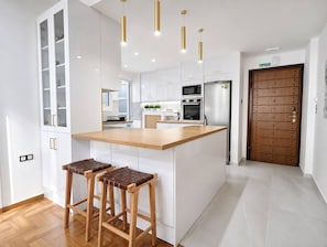 Eine moderne Küche mit weißen Schränken, einer Holzinsel, zwei Barhocker, Edelstahlgeräten und Pendelleuchten.