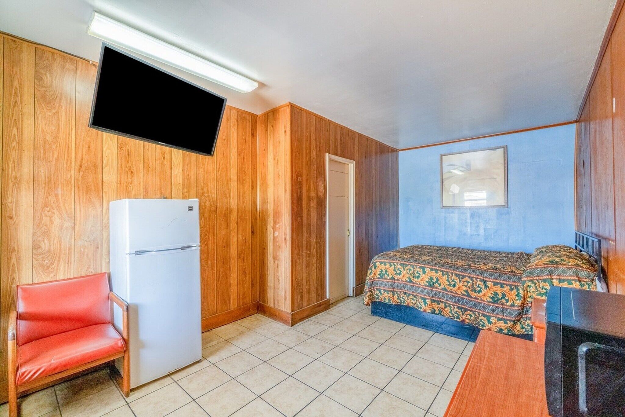 Ein gemütlicher Raum mit einem Bett, einem an der Wand befestigten Fernseher, einem Kühlschrank und einem Sessel.