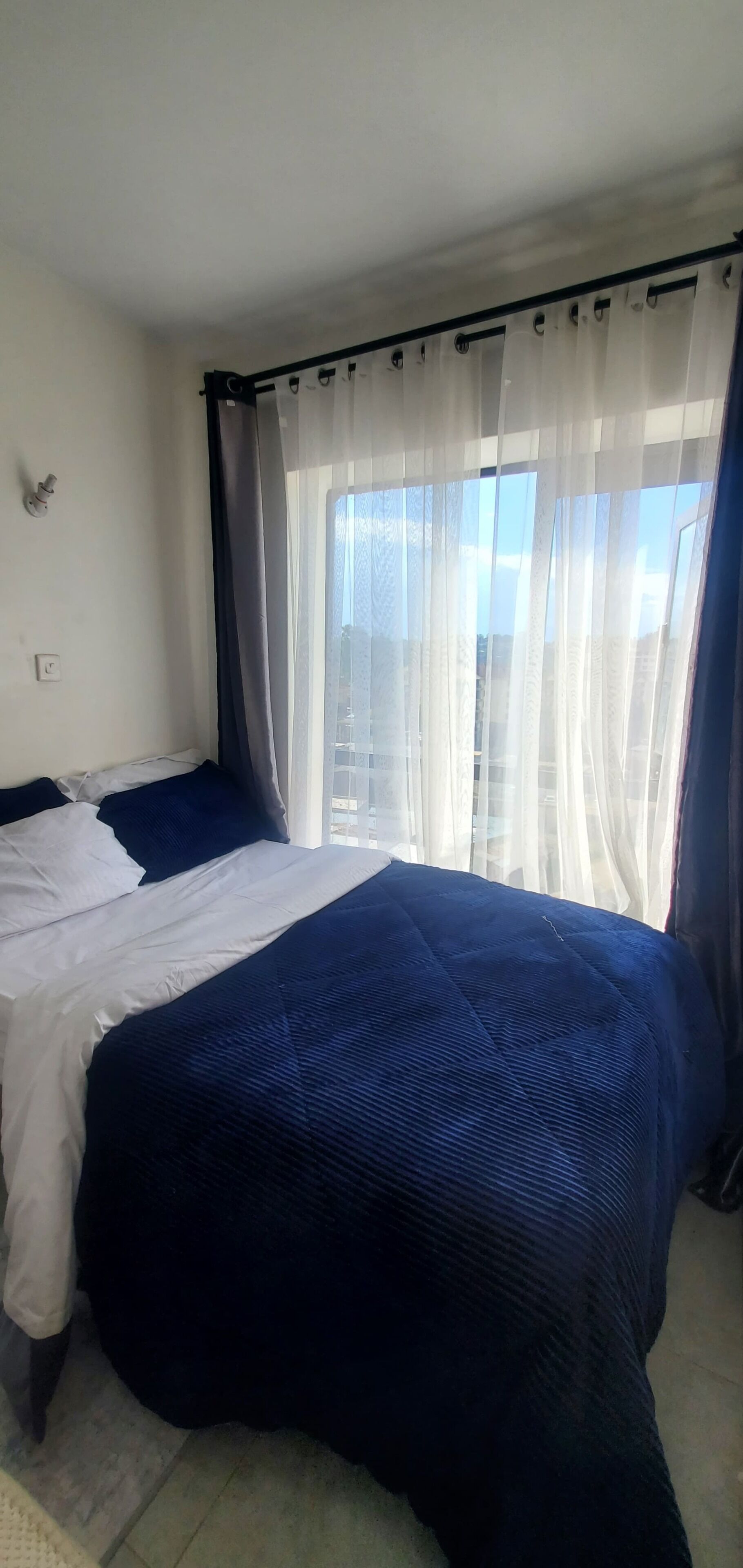 Et soveværelse med seng, gardiner, et vindue og en lampe på væggen.